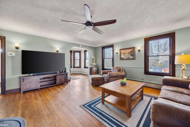 living room featuring arched walkways, a ceiling fan, light wood-style floors, a textured ceiling, and a baseboard heating unit