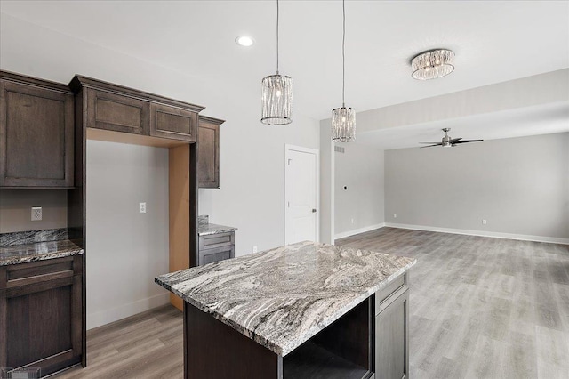 kitchen with light wood-style floors, ceiling fan with notable chandelier, dark brown cabinetry, a center island, and light stone counters