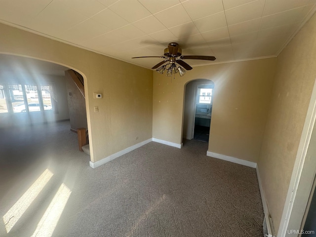 carpeted empty room featuring arched walkways, crown molding, a ceiling fan, and baseboards