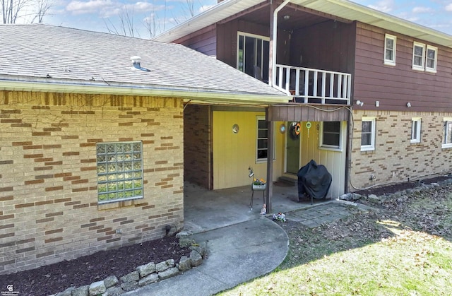 exterior space with roof with shingles, brick siding, a patio area, and a balcony
