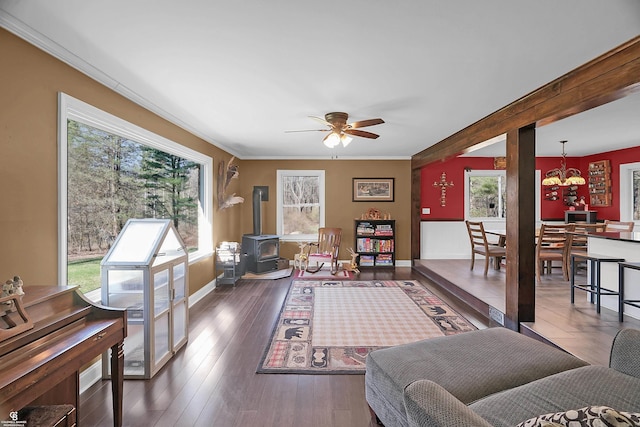 living room featuring wood finished floors, ceiling fan with notable chandelier, a wood stove, baseboards, and ornamental molding