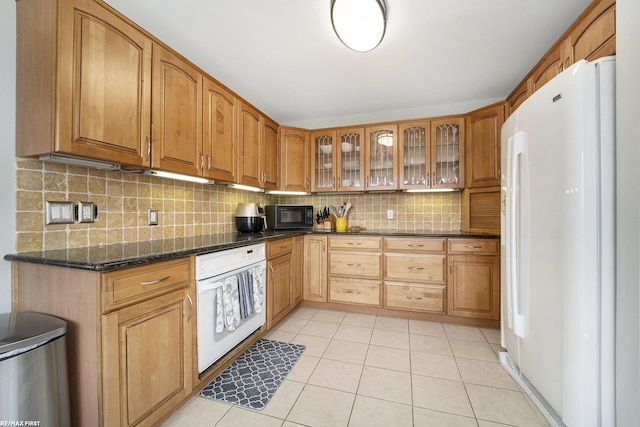 kitchen with dark stone countertops, light tile patterned floors, white appliances, glass insert cabinets, and tasteful backsplash
