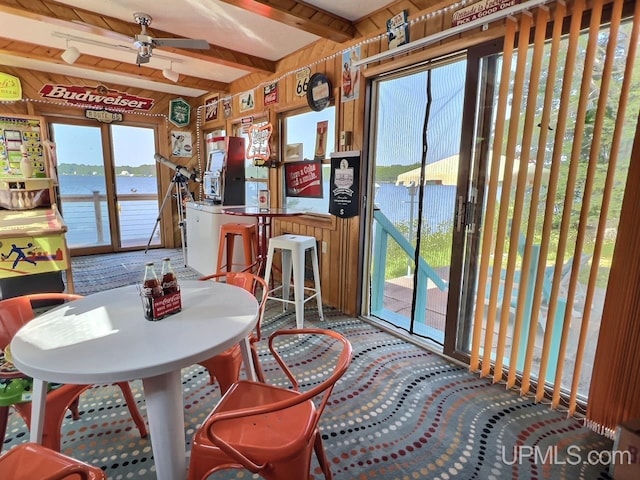 carpeted dining room featuring beamed ceiling, ceiling fan, wood walls, and a water view