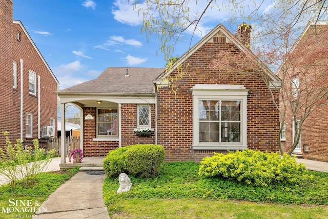 bungalow-style home with a chimney, ac unit, covered porch, brick siding, and a shingled roof