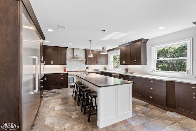 kitchen with a breakfast bar area, a kitchen island, dark brown cabinets, premium appliances, and wall chimney exhaust hood