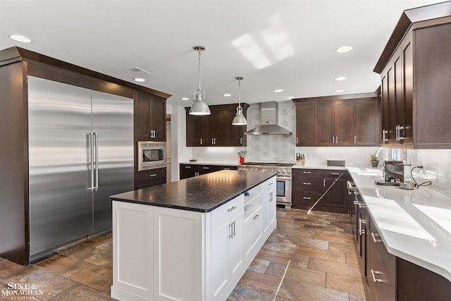 kitchen featuring wall chimney range hood, stone finish flooring, dark brown cabinets, built in appliances, and recessed lighting