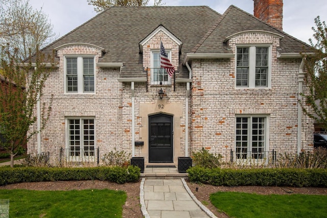 french country inspired facade featuring a shingled roof, a chimney, and brick siding