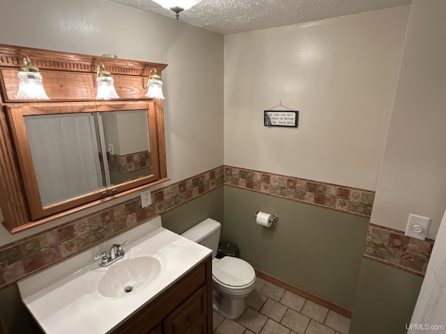 half bathroom featuring a textured ceiling, tile patterned flooring, vanity, a wainscoted wall, and toilet