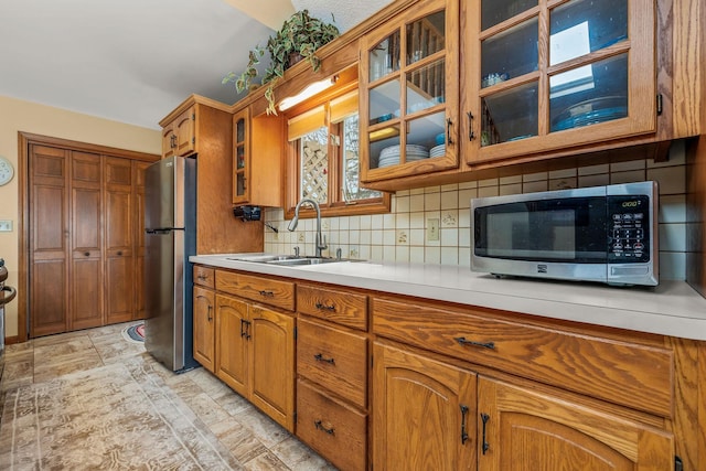 kitchen with brown cabinetry, appliances with stainless steel finishes, a sink, light countertops, and glass insert cabinets