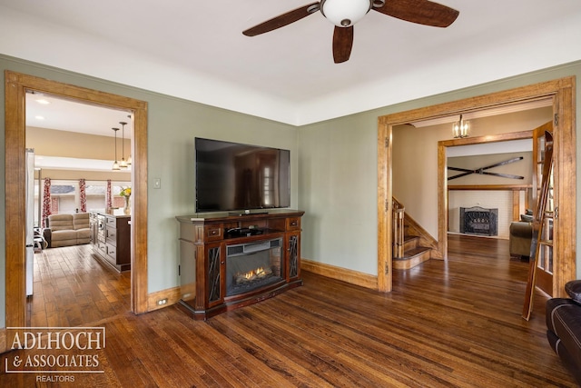 living room featuring a ceiling fan, a warm lit fireplace, baseboards, and dark wood-type flooring