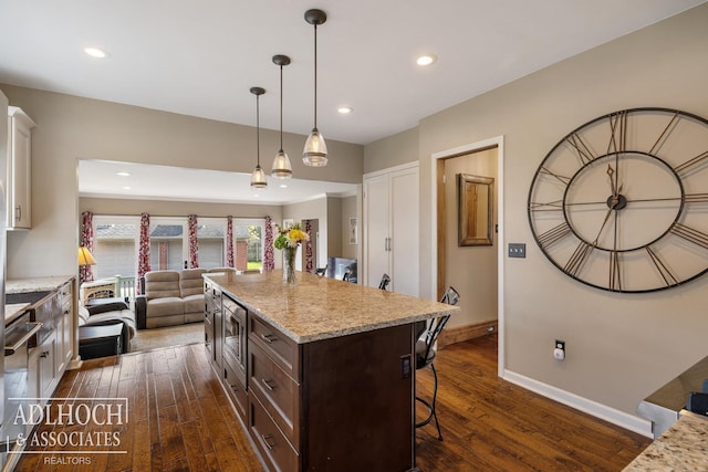 kitchen with a kitchen breakfast bar, dark brown cabinets, baseboards, stainless steel microwave, and dark wood-style floors