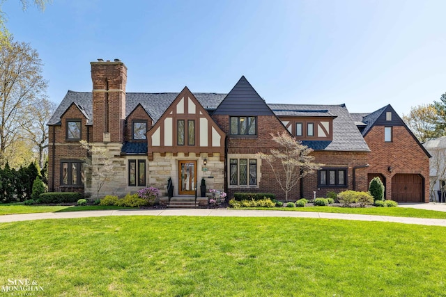 tudor-style house featuring a garage, a chimney, a shingled roof, and a front lawn