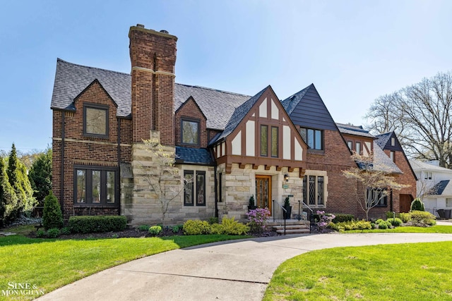 tudor home featuring roof with shingles, a chimney, a front lawn, and stone siding