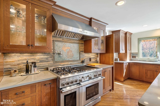 kitchen with light stone countertops, brown cabinetry, wall chimney exhaust hood, double oven range, and backsplash