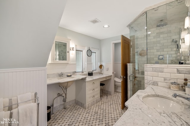 bathroom with visible vents, a shower stall, a wainscoted wall, two sinks, and toilet
