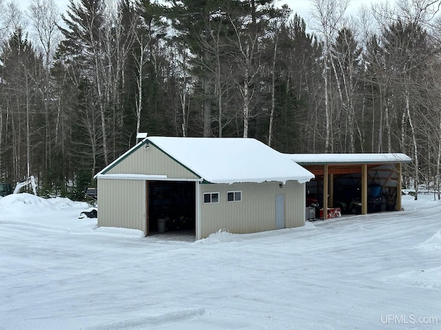 snow covered structure with an outdoor structure, an outbuilding, and a wooded view