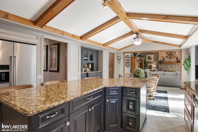 kitchen with light stone countertops, high end refrigerator, a ceiling fan, vaulted ceiling with beams, and a kitchen island