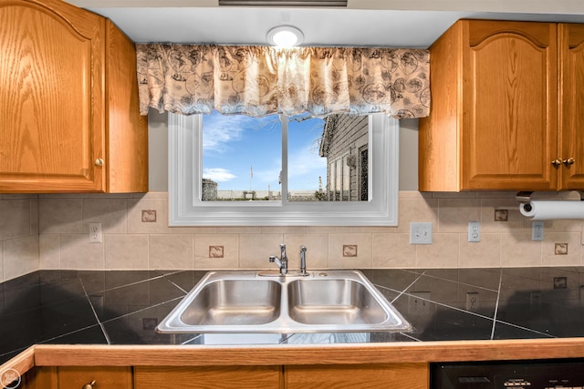 kitchen featuring brown cabinetry, dark countertops, dishwasher, a sink, and backsplash