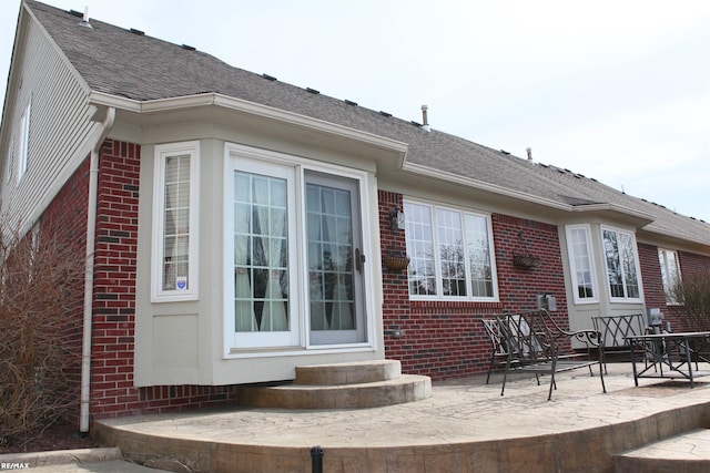 rear view of house with entry steps, a patio, roof with shingles, and brick siding