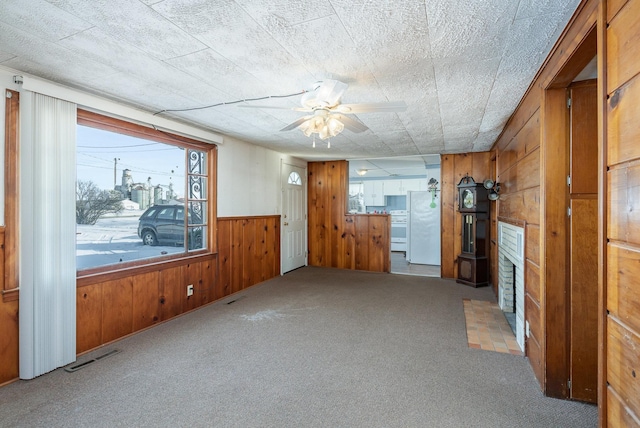 carpeted empty room featuring ceiling fan, visible vents, a fireplace with flush hearth, wainscoting, and wood walls