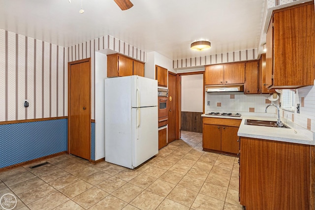 kitchen featuring brown cabinets, a sink, white appliances, light countertops, and wallpapered walls