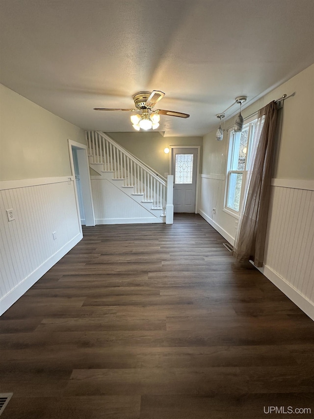 unfurnished living room with stairway, dark wood finished floors, wainscoting, and ceiling fan