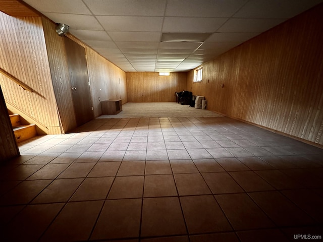 finished basement featuring a paneled ceiling, tile patterned flooring, wood walls, and stairway