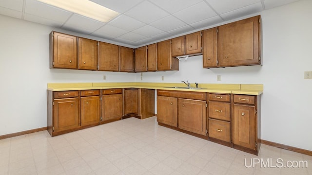kitchen featuring baseboards, a sink, a paneled ceiling, brown cabinets, and light countertops