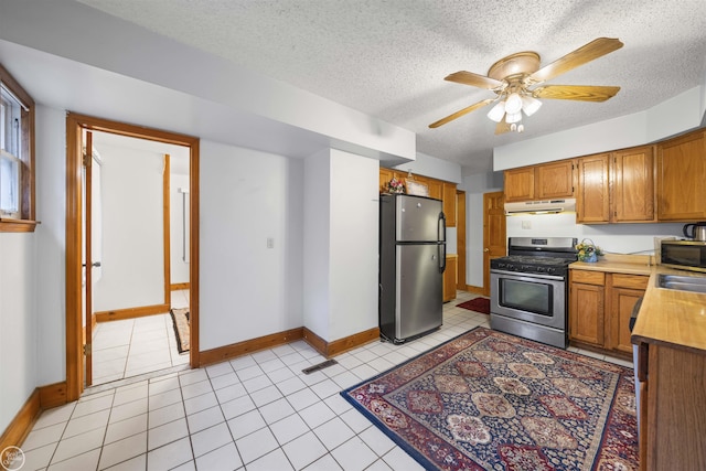 kitchen featuring appliances with stainless steel finishes, range hood, brown cabinets, baseboards, and a textured ceiling