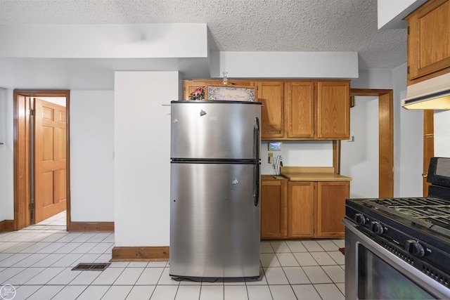 kitchen with stainless steel appliances, brown cabinets, light tile patterned floors, a textured ceiling, and under cabinet range hood