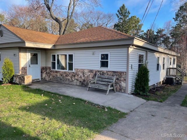rear view of property featuring a lawn, stone siding, central air condition unit, a shingled roof, and a patio area