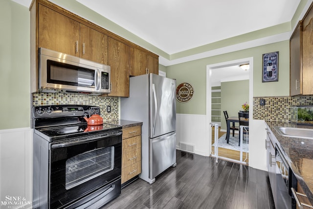 kitchen featuring stainless steel appliances, brown cabinetry, dark wood-type flooring, decorative backsplash, and wainscoting