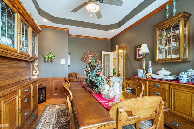 dining area with a ceiling fan, light wood finished floors, ornamental molding, a raised ceiling, and wainscoting