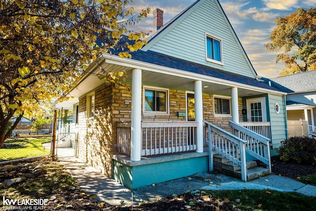 bungalow-style house with a porch, a shingled roof, a chimney, stone siding, and fence