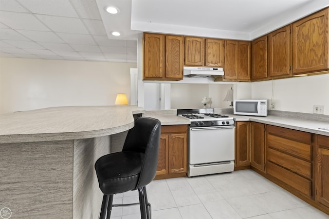 kitchen featuring white appliances, under cabinet range hood, brown cabinets, light countertops, and recessed lighting