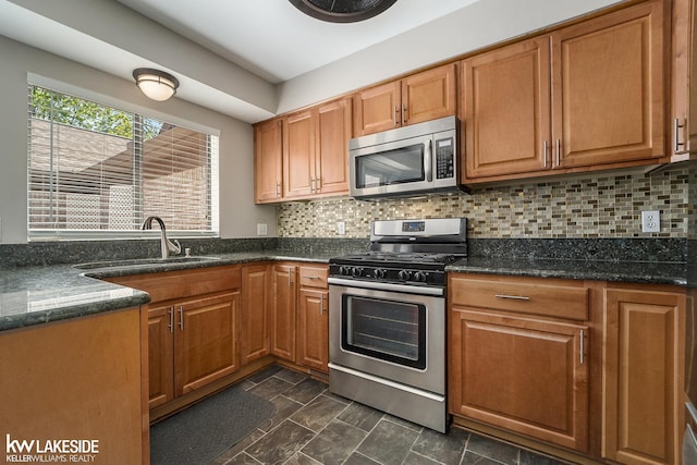 kitchen featuring stainless steel appliances, brown cabinetry, backsplash, a sink, and dark stone counters