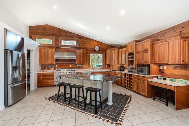 kitchen with appliances with stainless steel finishes, brown cabinets, wall chimney range hood, a center island with sink, and a breakfast bar