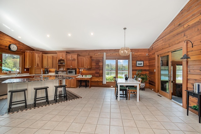 kitchen with vaulted ceiling, wooden walls, stainless steel microwave, brown cabinetry, and an island with sink