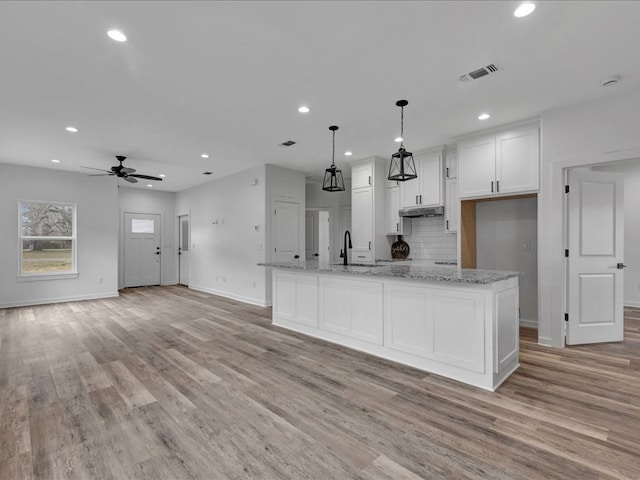 kitchen with light stone counters, hanging light fixtures, a center island with sink, and white cabinets