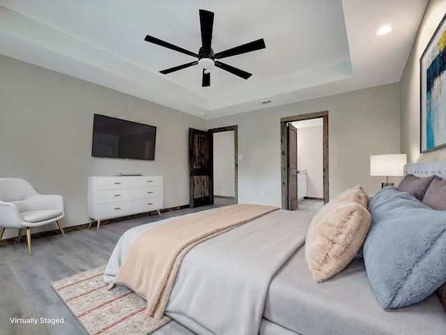 bedroom featuring a tray ceiling, ceiling fan, ensuite bathroom, and light wood-type flooring