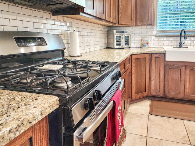 kitchen with light tile patterned floors, a sink, decorative backsplash, stainless steel gas stove, and brown cabinets