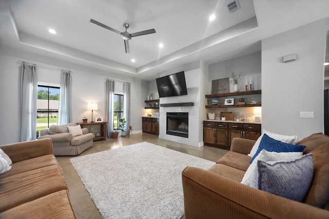 living room featuring ceiling fan, a fireplace, a tray ceiling, and finished concrete floors