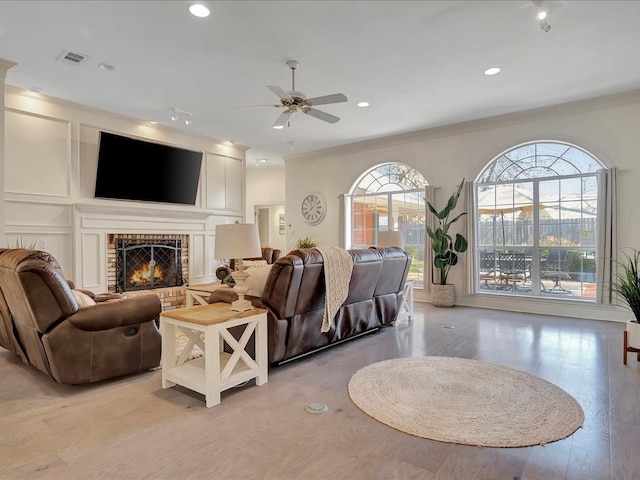 living room featuring ceiling fan, light wood-type flooring, ornamental molding, and a brick fireplace