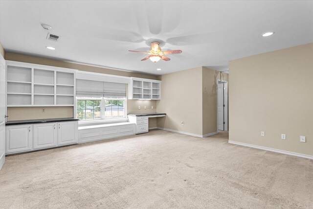 unfurnished living room with ceiling fan, built in desk, and light carpet