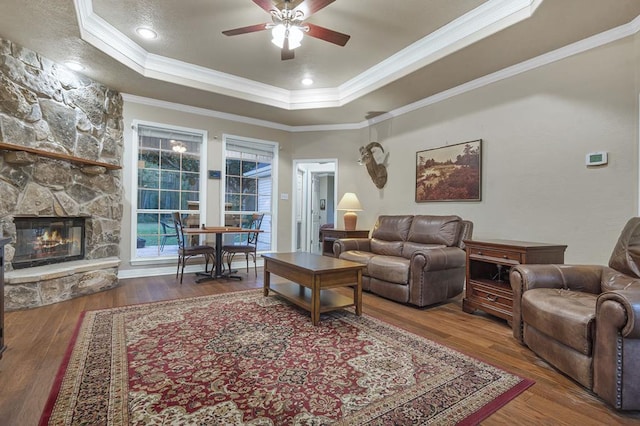 living room with a raised ceiling, ceiling fan, crown molding, wood-type flooring, and a stone fireplace