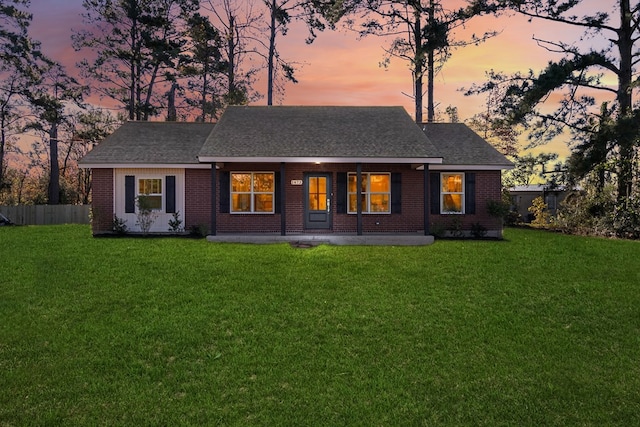 back of house at dusk with a shingled roof, brick siding, a yard, and fence