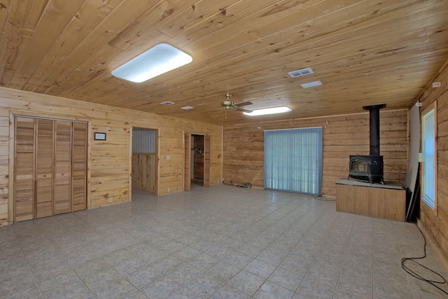 unfurnished living room featuring visible vents, a wood stove, ceiling fan, wood walls, and wooden ceiling