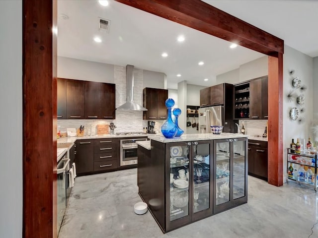 kitchen with wall chimney range hood, decorative backsplash, dark brown cabinetry, a kitchen island with sink, and stainless steel appliances