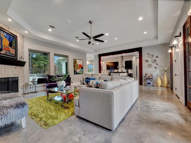 living room featuring ceiling fan, a raised ceiling, and a stone fireplace