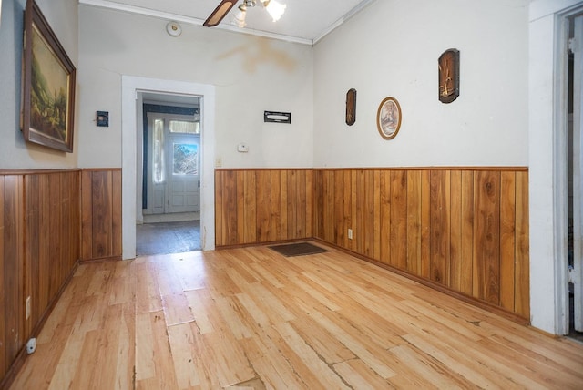 spare room with crown molding, light wood-style flooring, a ceiling fan, wainscoting, and wooden walls